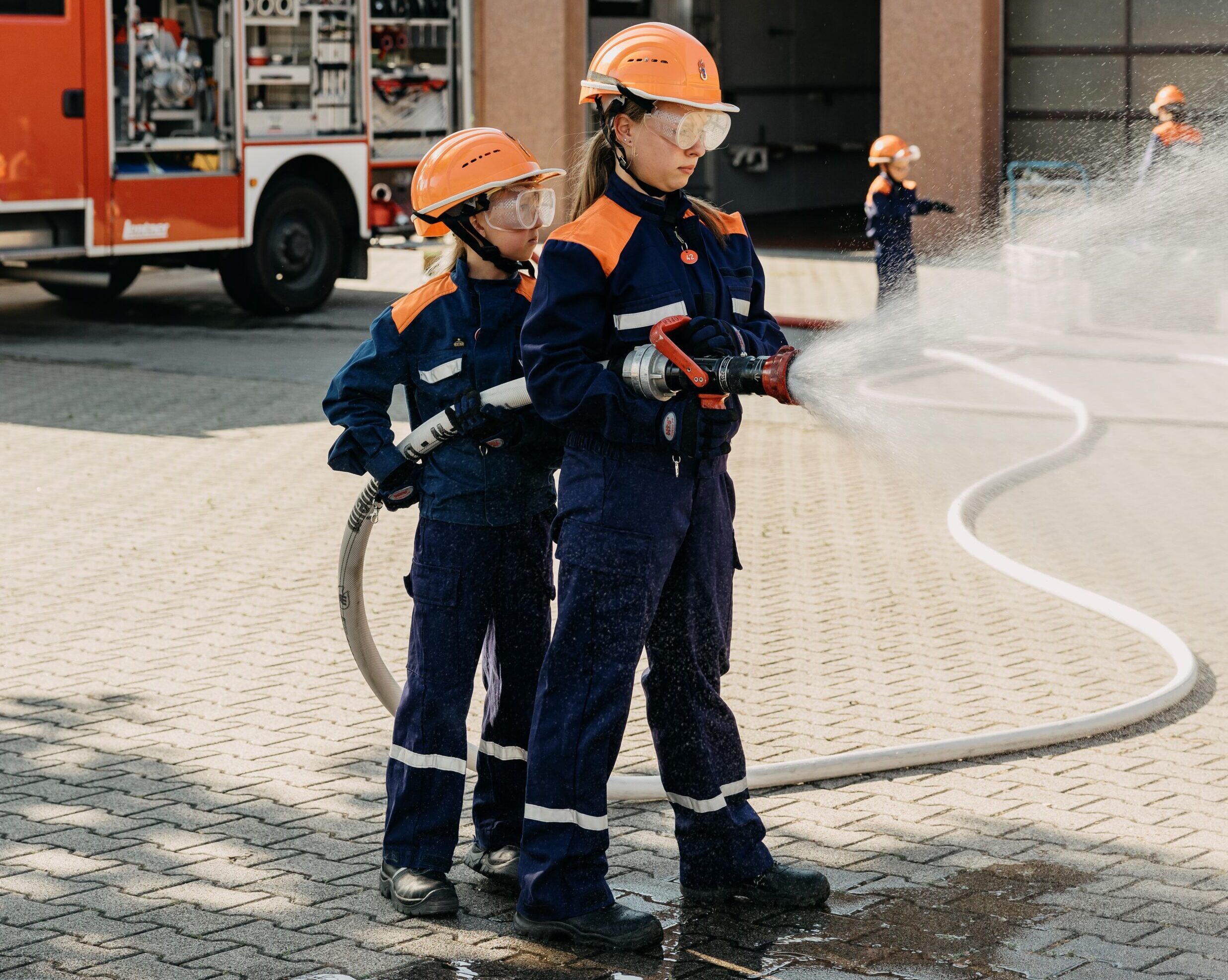 Zwei Kinder in Feuerwehrmontur spritzen mit einem Schlauch, als würden sie ein Feuer löschen.
